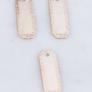 May include: Three gold-colored metal tags with an ornate design. The tags are rectangular and have a hole at the top for hanging.