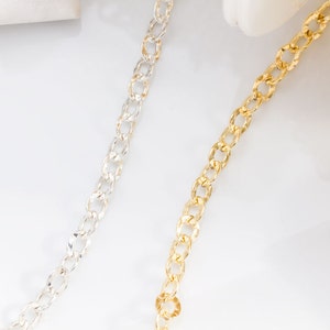 May include: Two chains, one silver and one gold, are laid out on a white surface. Both chains are made of a curb link style.