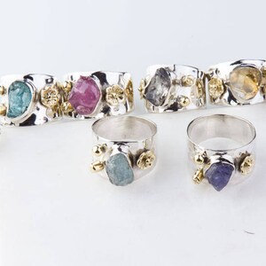 May include: A set of silver rings with different colored gemstones. Each ring features a unique, rough-cut gemstone set in a silver band with gold accents. The gemstones include aquamarine, amethyst, topaz, and pink tourmaline.