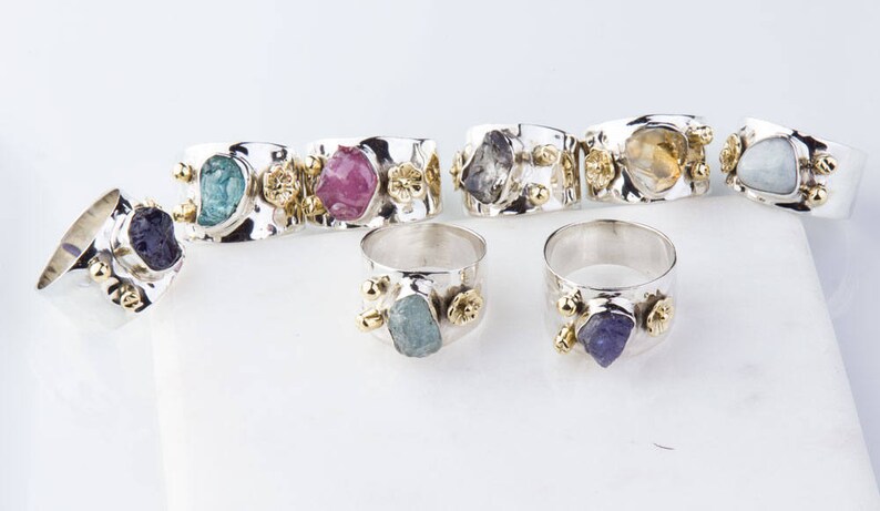 May include: A set of silver rings with different colored gemstones. Each ring features a unique, rough-cut gemstone set in a silver band with gold accents. The gemstones include aquamarine, amethyst, topaz, and pink tourmaline.