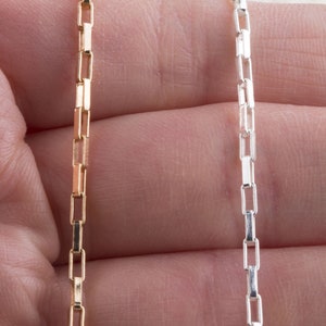 May include: Two chains, one gold and one silver, with a box link design. The chains are held in a hand.