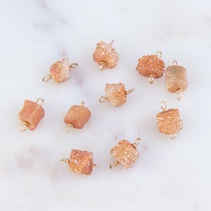May include: Ten peach-colored crystal beads with gold jump rings. The beads are square shaped and have a rough, textured surface.