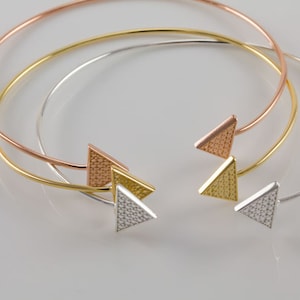 May include: Three gold bangles with triangle-shaped charms. The bangles are in rose gold, yellow gold, and silver.