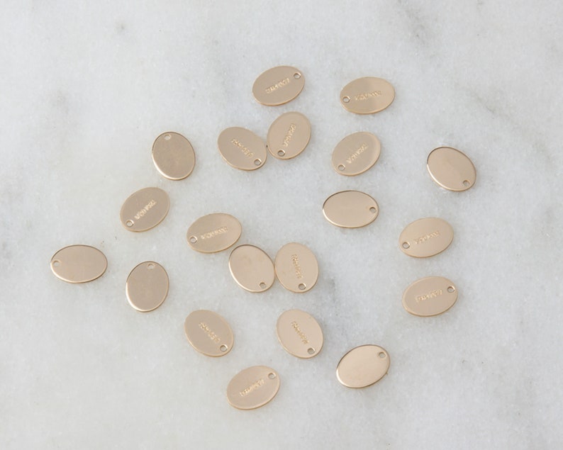 May include: A collection of 18 gold oval-shaped metal tags with various words engraved on them. The tags have a hole at the top for attaching to a necklace or bracelet.