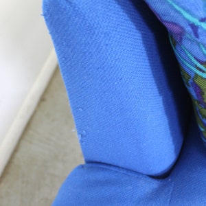 May include: Close-up of a blue fabric armchair with a floral patterned pillow. The fabric has a textured weave.