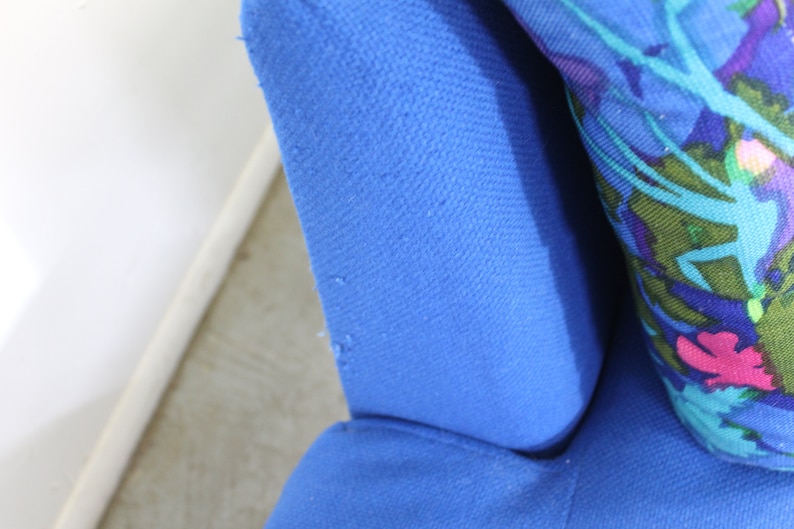 May include: Close-up of a blue fabric armchair with a floral patterned pillow. The fabric has a textured weave.