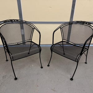 May include: Two black metal chairs with a mesh design. The chairs have a curved back and arms. The chairs are sitting on a gray concrete floor.