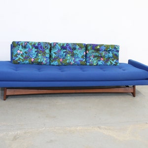 May include: A blue sofa with a wooden frame and three floral patterned pillows. The sofa is sitting on a concrete floor.