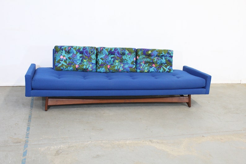 May include: A blue sofa with a wooden frame and three floral patterned pillows. The sofa is sitting on a concrete floor.