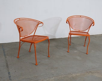 May include: Two orange metal chairs with curved backs and seats made of a mesh pattern. The chairs have slender legs and are set against a white wall and gray floor. The chairs are a bright, solid color.