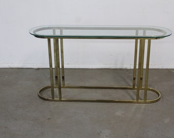 May include: A gold metal console table with a clear glass top. The table has a rectangular shape with rounded edges and a simple design.