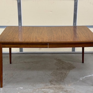 May include: A rectangular wooden dining table with a brown finish and a leaf extension. The table has four legs and a simple design.