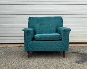 May include: Teal armchair with a patterned fabric design. The chair has a rectangular shape with a seat cushion and armrests. The wooden legs are dark brown. The background is a white garage door.