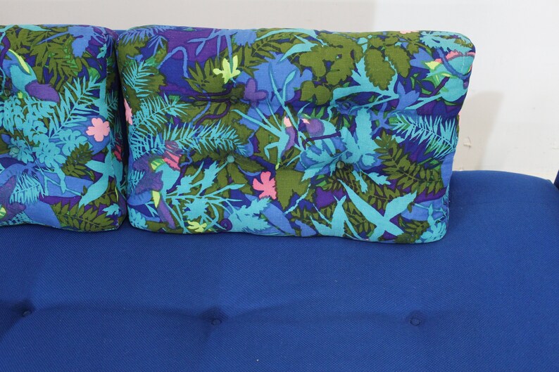 May include: A blue sofa with two floral patterned pillows. The pillows have a blue, green, and pink floral design.