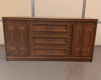 May include: A mid-century modern dresser with three drawers and two cabinet doors. The dresser is made of wood and has a dark brown finish. The doors have a decorative design with oval cutouts.