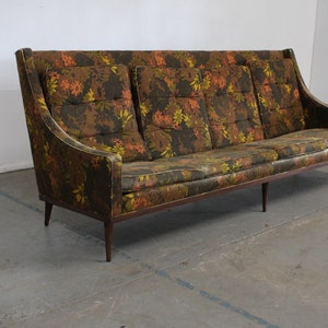 May include: A long, brown sofa with a floral patterned upholstery. The sofa has a high back and a curved armrest. The sofa is sitting on a concrete floor.