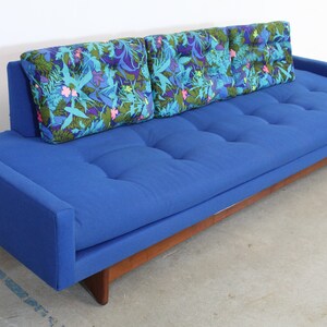 May include: A blue tufted sofa with a wooden base and three floral patterned pillows.