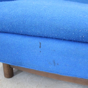 May include: Close-up of a blue fabric sofa with wooden legs. The fabric has a slight texture and there are a few dark marks on the surface.