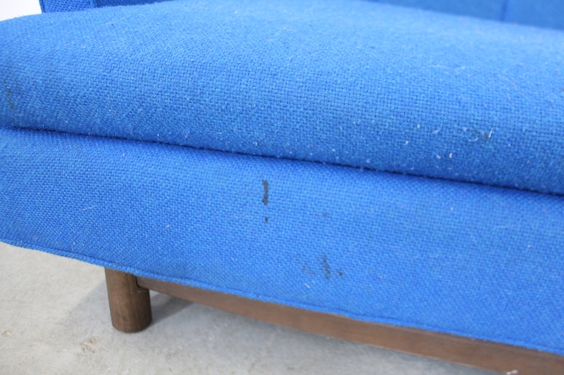May include: Close-up of a blue fabric sofa with wooden legs. The fabric has a slight texture and there are a few dark marks on the surface.