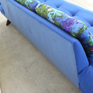 May include: A blue sofa with a floral patterned pillow. The sofa has a tufted back and a slightly curved armrest.