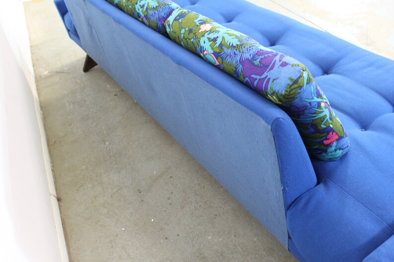 May include: A blue sofa with a floral patterned pillow. The sofa has a tufted back and a slightly curved armrest.