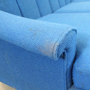 May include: A close-up of a blue fabric sofa armrest. The fabric has a textured weave and shows signs of wear.