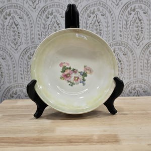 May include: A vintage, cream-colored ceramic bowl with a floral design in the center. The flowers are pink, white, and blue. The bowl is displayed on a black stand against a patterned wallpaper background. The bowl has a diameter of approximately 10 inches.