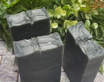 May include: Three bars of charcoal soap, each with a slightly different texture, are arranged on a stone-like surface. The soap is a dark gray color.