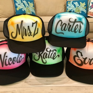 May include: Five black mesh trucker hats with different colored fronts and names written in black cursive. The names are "Mrs. Z", "Carter", "Nicole", "Katie", and "Eva".