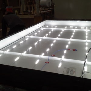 Puede incluir: Una caja de luz rectangular blanca con un marco negro y varias filas de luces LED. Las luces est&aacute;n conectadas por cables y son visibles a trav&eacute;s de la cubierta de pl&aacute;stico transparente.