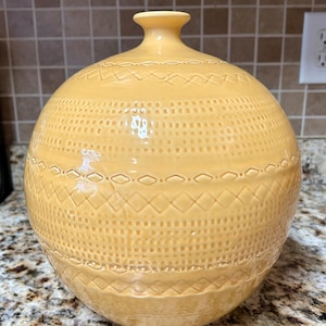 May include: A spherical ceramic vase in a soft yellow hue, featuring a narrow neck and a small circular opening. The exterior surface is decorated with a repeating embossed pattern of horizontal bands, including geometric diamonds and dotted textures.