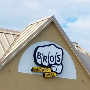 Puede incluir: Un letrero blanco con un logotipo de pu&ntilde;o negro que dice "Bros Sandwich Shack". El letrero est&aacute; en el lateral de un edificio con un techo beige.