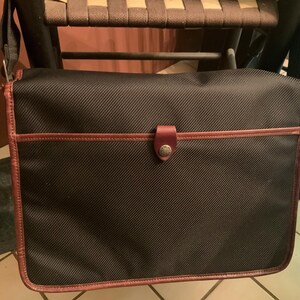Pode incluir: Uma bolsa tiracolo preta com textura de tecido diagonal, apresentando acabamento em couro marrom nas bordas e um fecho de aba central preso por uma aba de couro marrom com um bot&atilde;o met&aacute;lico. A bolsa inclui uma al&ccedil;a de ombro ajust&aacute;vel.