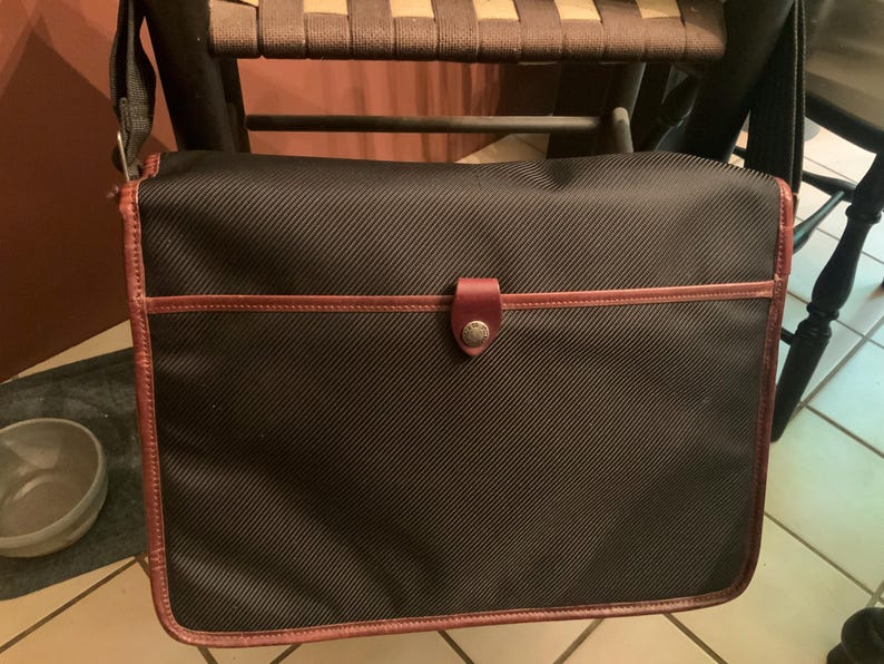 Pode incluir: Uma bolsa tiracolo preta com textura de tecido diagonal, apresentando acabamento em couro marrom nas bordas e um fecho de aba central preso por uma aba de couro marrom com um bot&atilde;o met&aacute;lico. A bolsa inclui uma al&ccedil;a de ombro ajust&aacute;vel.