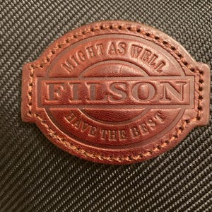 Pode incluir: Um emblema de couro castanho com bordas costuradas fixado num tecido preto texturizado. O emblema apresenta o texto "FILSON" em relevo no centro, com a frase "MIGHT AS WELL HAVE THE BEST" arqueada em torno da parte superior e inferior.