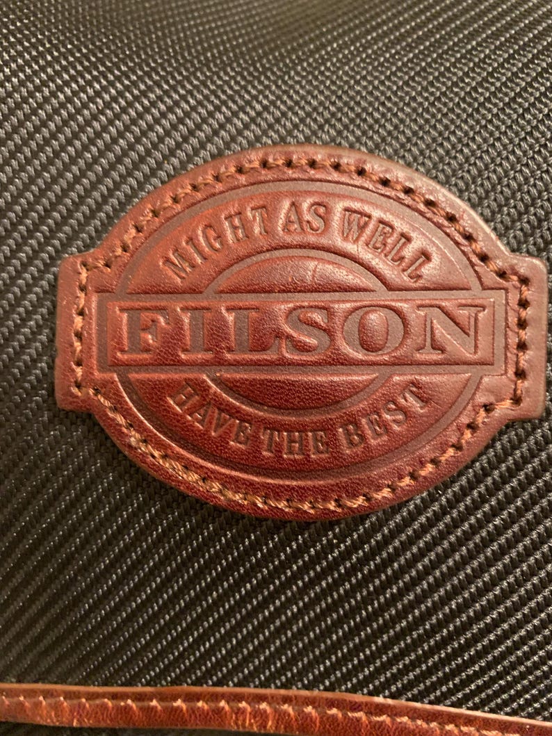 Pode incluir: Um emblema de couro castanho com bordas costuradas fixado num tecido preto texturizado. O emblema apresenta o texto "FILSON" em relevo no centro, com a frase "MIGHT AS WELL HAVE THE BEST" arqueada em torno da parte superior e inferior.