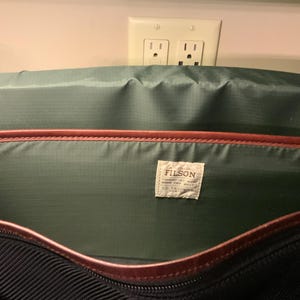 Pode incluir: O interior de uma bolsa com forro de nylon verde escuro e acabamento em couro marrom nas bordas. Uma etiqueta de tecido retangular branca est&aacute; costurada no painel traseiro com o texto FILSON impresso em preto e texto adicional.