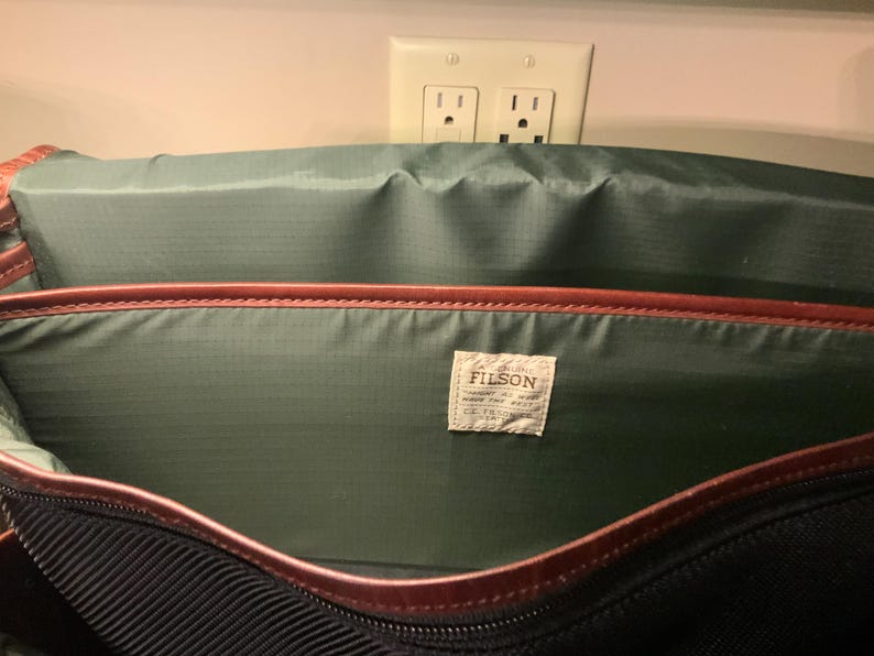 Pode incluir: O interior de uma bolsa com forro de nylon verde escuro e acabamento em couro marrom nas bordas. Uma etiqueta de tecido retangular branca est&aacute; costurada no painel traseiro com o texto FILSON impresso em preto e texto adicional.