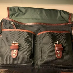 Pode incluir: Uma bolsa mensageiro verde-escura com exterior em tecido texturizado, uma aba grande e dois bolsos frontais fixados com tiras de couro marrom e fivelas de metal. A bolsa inclui um compartimento com z&iacute;per e detalhes de acabamento em couro marrom.