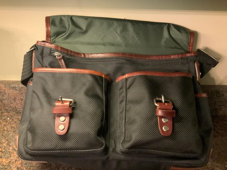 Pode incluir: Uma bolsa mensageiro verde-escura com exterior em tecido texturizado, uma aba grande e dois bolsos frontais fixados com tiras de couro marrom e fivelas de metal. A bolsa inclui um compartimento com z&iacute;per e detalhes de acabamento em couro marrom.
