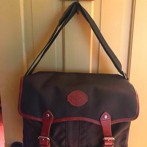 Pode incluir: Uma bolsa carteiro preta com corpo de tecido texturizado, aba superior com duas tiras de couro marrom, fivelas de metal prateado e um emblema circular de couro marrom com o texto Filson em relevo no centro da aba.