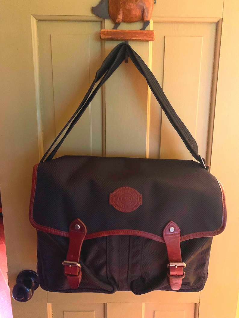Pode incluir: Uma bolsa carteiro preta com corpo de tecido texturizado, aba superior com duas tiras de couro marrom, fivelas de metal prateado e um emblema circular de couro marrom com o texto Filson em relevo no centro da aba.
