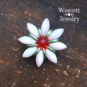 May include: A handcrafted flower-shaped jewelry piece. It features white, petal-like beads, green ring accents, and a red center. The jewelry is set against a dark wooden background. The text "Wescott Jewelry" is visible.