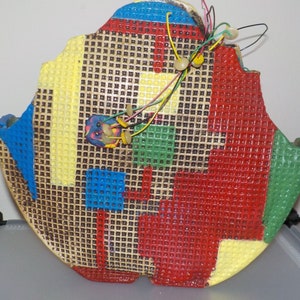 May include: A colorful, grid-like, circular object with a damaged edge. The object is made of a textured material and is painted in shades of red, yellow, blue, and green. There are several wires and small, colorful objects attached to the object.