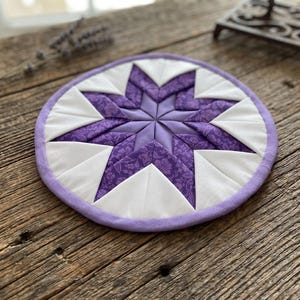 May include: A round, quilted trivet with a star design. The star is made of purple fabric with a floral pattern, surrounded by white triangles and a lavender border. The trivet is on a wooden surface.