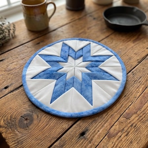 May include: A round, quilted trivet with a starburst design. The star is made of white and blue fabric, with a blue border. The trivet is on a wooden table. Kitchen decor.