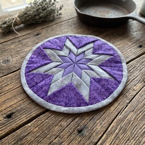 May include: A round, quilted trivet with a starburst design. The trivet features a central star in shades of purple and gray, with a floral pattern on the purple sections. The edge is finished with a gray binding. It rests on a rustic wooden surface.