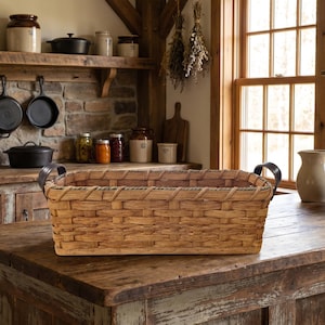May include: A rectangular woven basket with dark handles sits on a rustic wooden countertop. The basket is light brown and has a textured appearance. The kitchen setting includes shelves with jars and cast iron pans, suggesting a farmhouse theme.