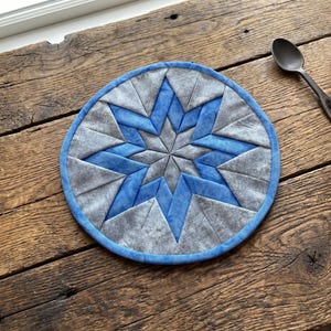May include: A round, quilted trivet with a starburst design. The trivet features a central star in shades of blue and gray, surrounded by a blue border. It rests on a rustic wooden surface, with a spoon and mug nearby.