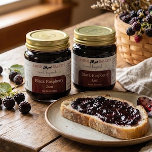 May include: Two jars of black raspberry jam with gold lids sit on a wooden table, alongside a slice of bread spread with jam. Fresh raspberries and a basket of berries are also visible. The jars are labeled "Amish Baskets and beyond."